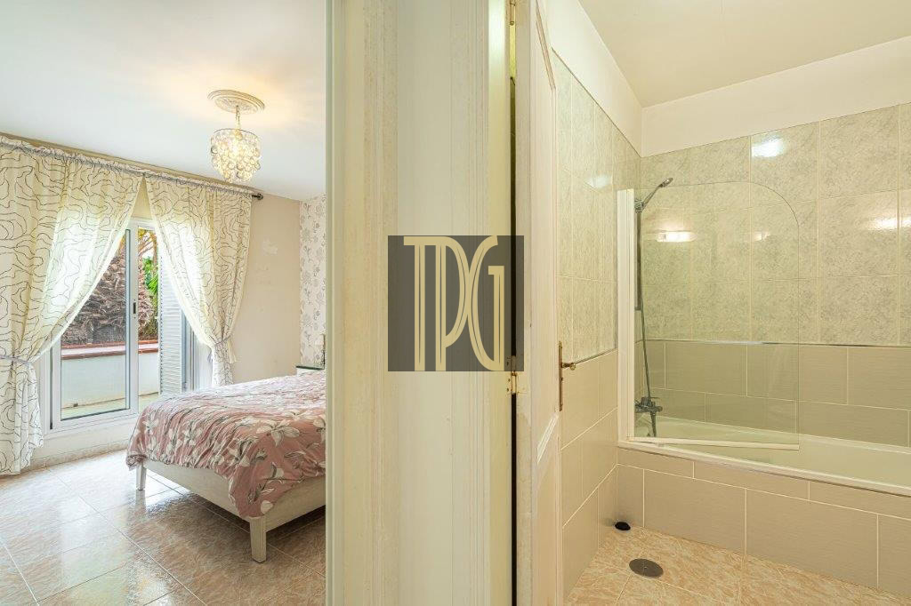 Bright bedroom with a pink floral bedspread, sheer curtains, and a balcony door on the left, adjacent to a tiled bathroom with a bathtub on the right