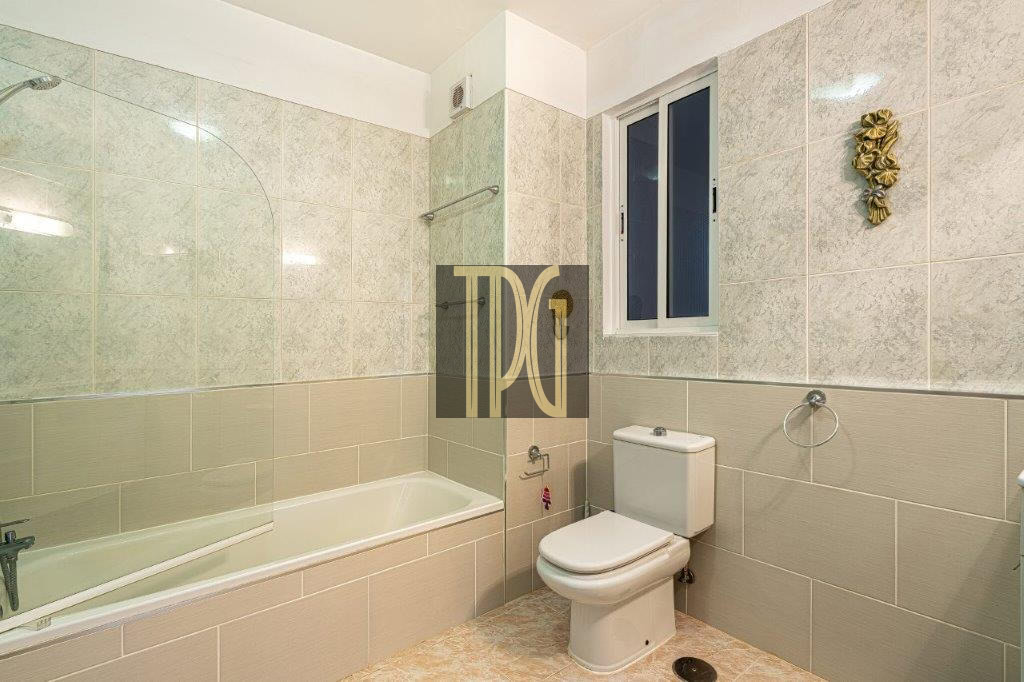 Bathroom with a white bathtub and glass shower panel, a toilet, and beige tiled walls with a small window above the toilet.