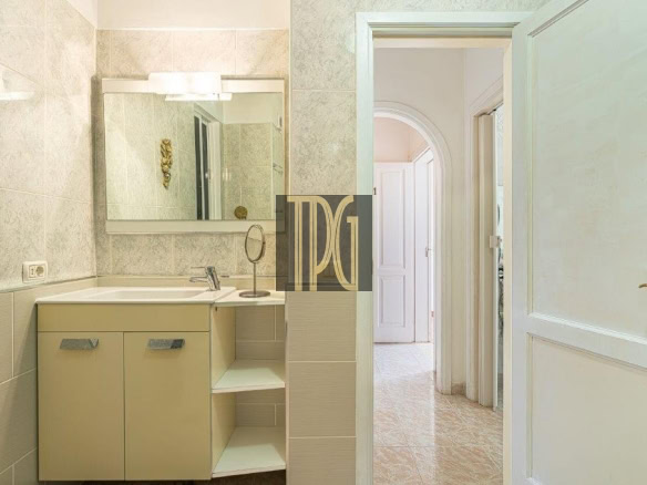 A beige bathroom vanity with a sink and a large wall mirror, tiled walls, and an open doorway leading to a bright hallway.
