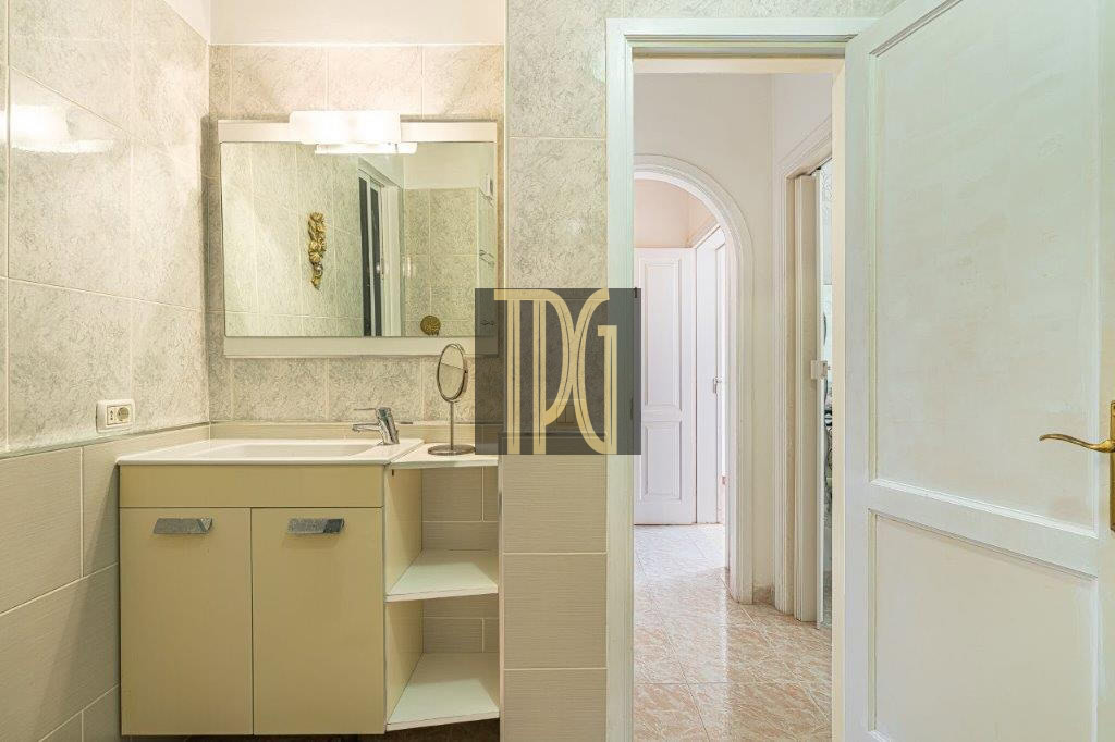 A beige bathroom vanity with a sink and a large wall mirror, tiled walls, and an open doorway leading to a bright hallway.