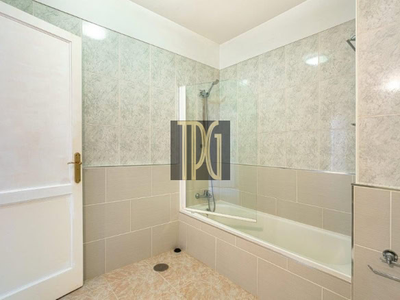 Bathroom with a white tub and curved glass shower screen, gray tiled walls, beige floor tiles, and a white door on the left.