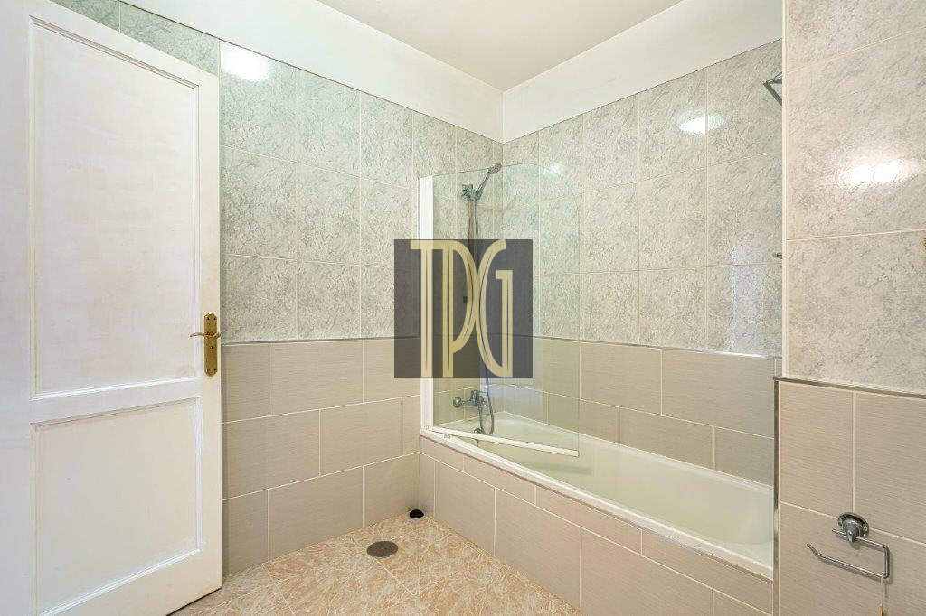 Bathroom with a white tub and curved glass shower screen, gray tiled walls, beige floor tiles, and a white door on the left.