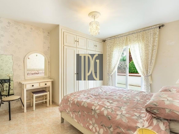 Bright bedroom with two twin beds (or a double) in pink floral bedding, a white vanity with mirror, and a large built-in wardrobe near a sliding glass door with patterned curtains.