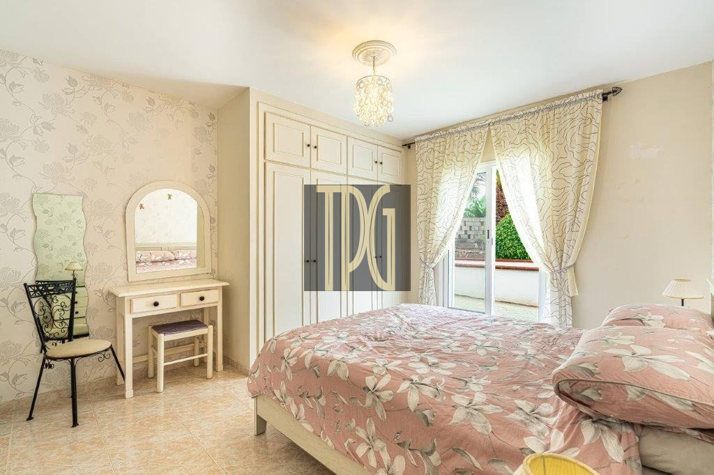 Bright bedroom with two twin beds (or a double) in pink floral bedding, a white vanity with mirror, and a large built-in wardrobe near a sliding glass door with patterned curtains.
