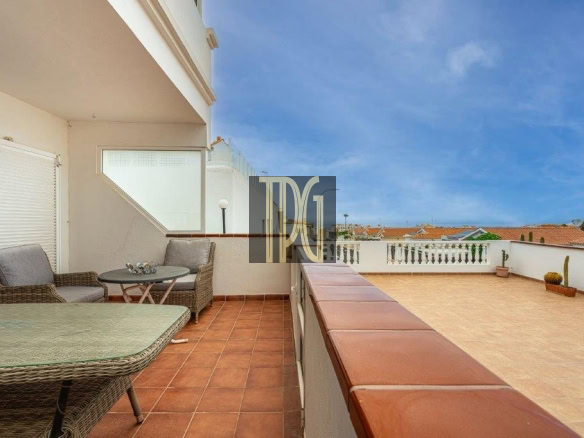 Terrace with wicker seating and a glass table on terracotta tiles, overlooking a white railing and blue sky.