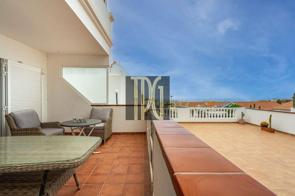 Terrace with wicker seating and a glass table on terracotta tiles, overlooking a white railing and blue sky.