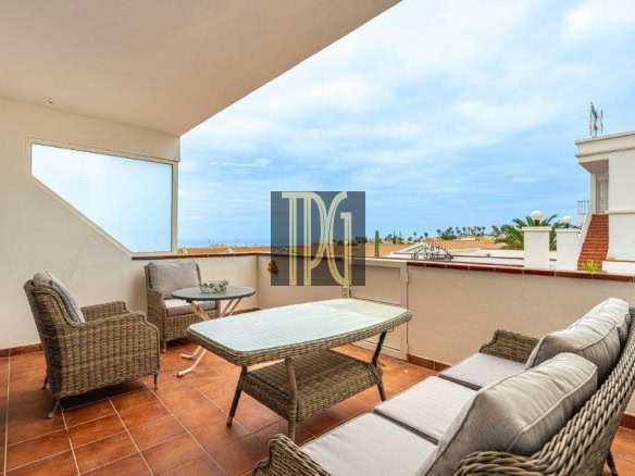Balcony seating area with wicker chairs, a glass table, and a sofa, overlooking the sea and nearby buildings.