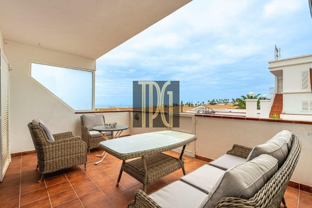 Balcony seating area with wicker chairs, a glass table, and a sofa, overlooking the sea and nearby buildings.