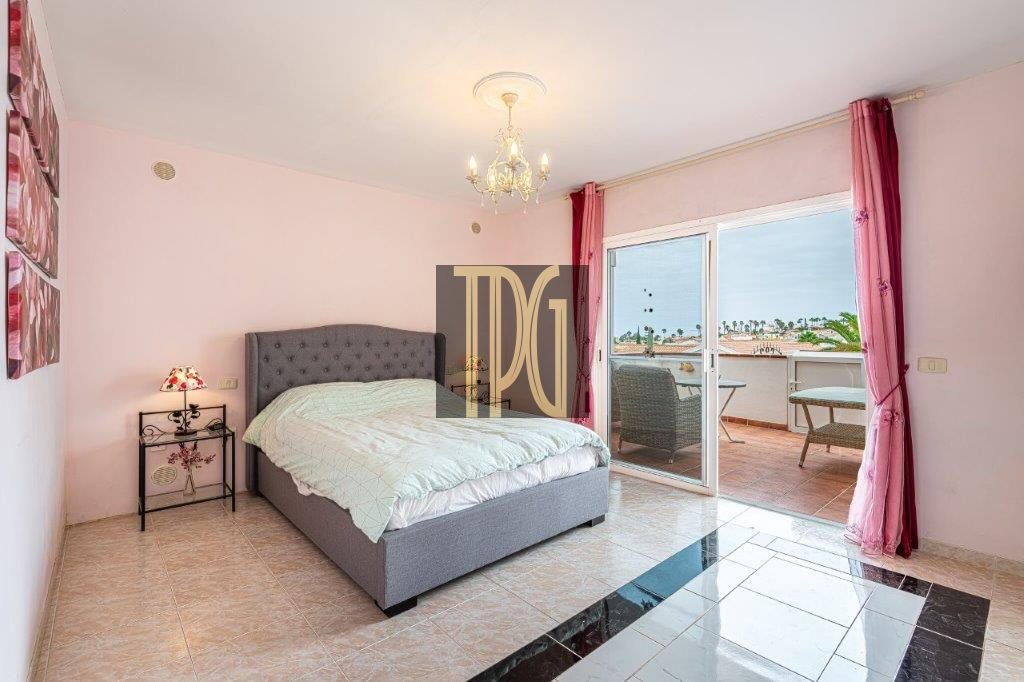Bedroom with pink walls, tufted gray bed, mint quilt, and balcony doors to a seating area with an ocean view in the distance.