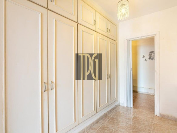 Long row of cream built-in wardrobes along a narrow hallway with tiled floor and a doorway to another room on the right, bright lighting above.