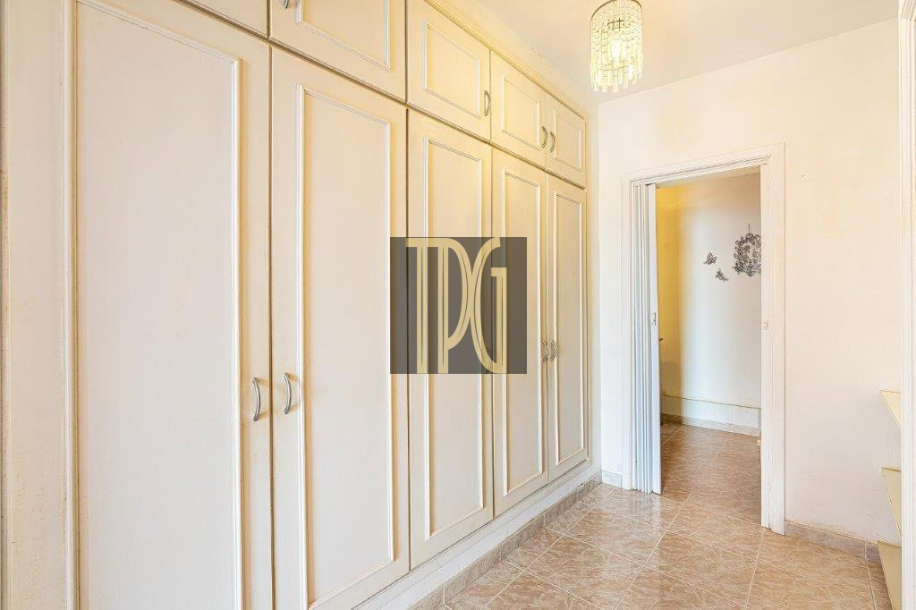 Long row of cream built-in wardrobes along a narrow hallway with tiled floor and a doorway to another room on the right, bright lighting above.