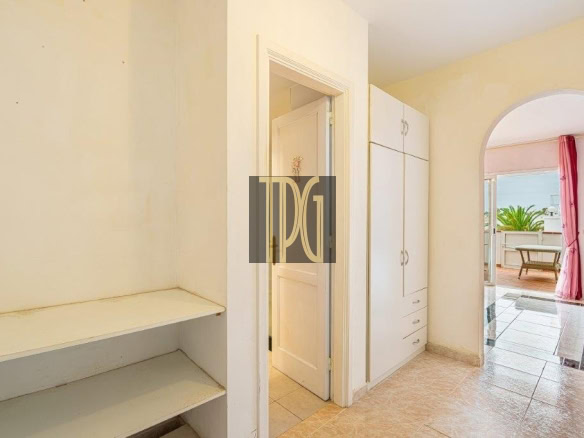 Bright hallway with built-in white wardrobes and open door, plus a shelving unit on the left; arched doorway to a pink living area with a balcony beyond.