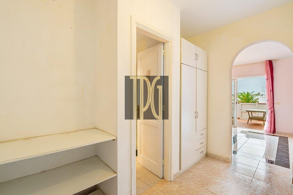 Bright hallway with built-in white wardrobes and open door, plus a shelving unit on the left; arched doorway to a pink living area with a balcony beyond.