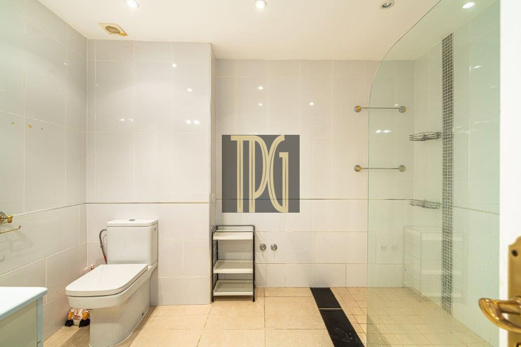 Modern white-tiled bathroom featuring a toilet, a small gray shelving cart, and a glass-enclosed shower on the right; beige tiled floor and wall-mounted towel racks create a clean, minimalist look.