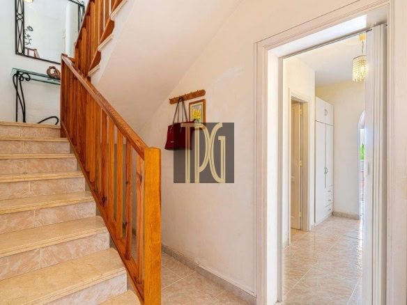 Beige-tiled staircase with a wooden handrail running along a white-walled hallway, leading to an upper floor.
