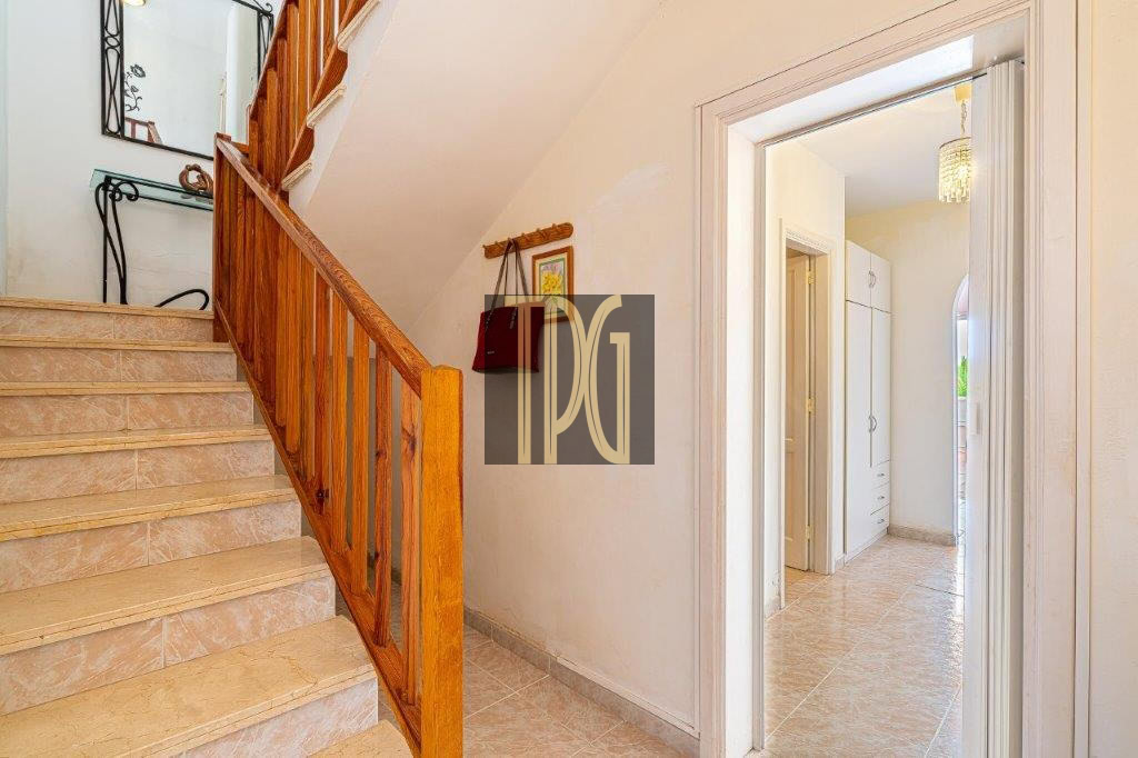 Beige-tiled staircase with a wooden handrail running along a white-walled hallway, leading to an upper floor.