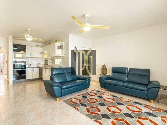 Open-concept living room with two teal leather couches, a geometric rug, and a white kitchen in the background on tiled floors.