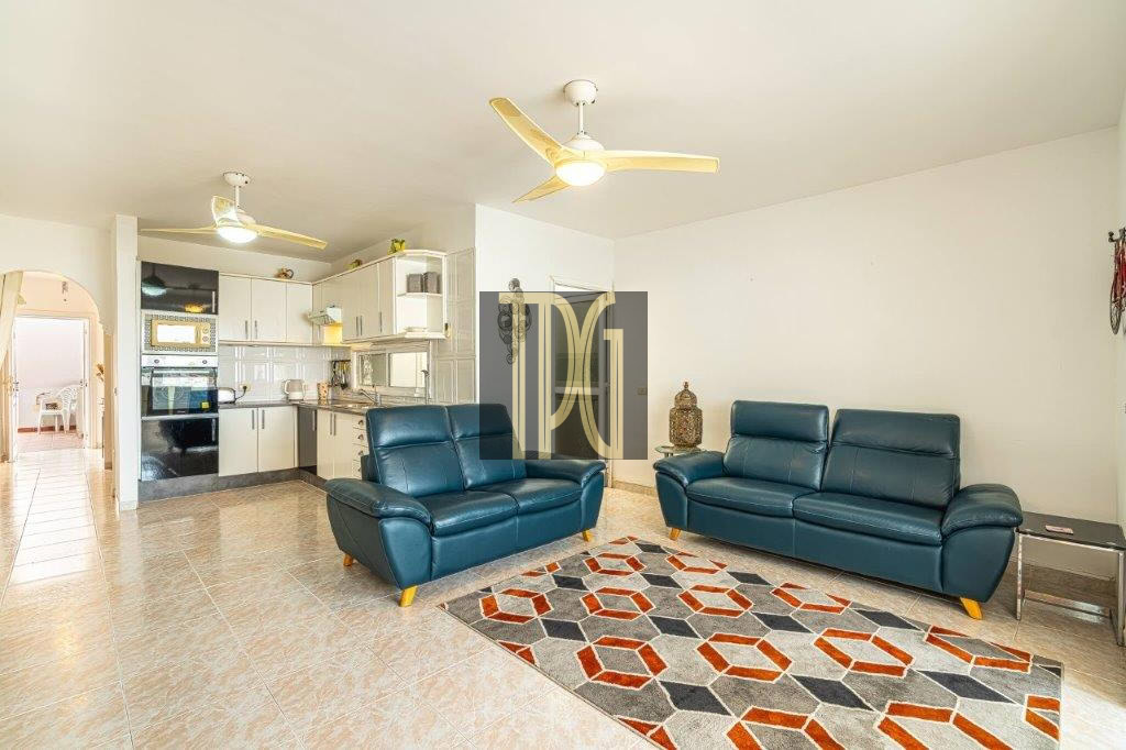Open-concept living room with two teal leather couches, a geometric rug, and a white kitchen in the background on tiled floors.