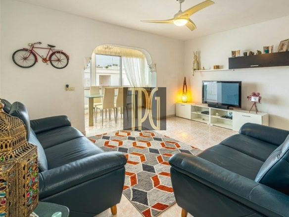 Living room with two blue leather couches facing a TV on a white console and a colorful geometric rug in a bright open space with an arched doorway to the dining area.