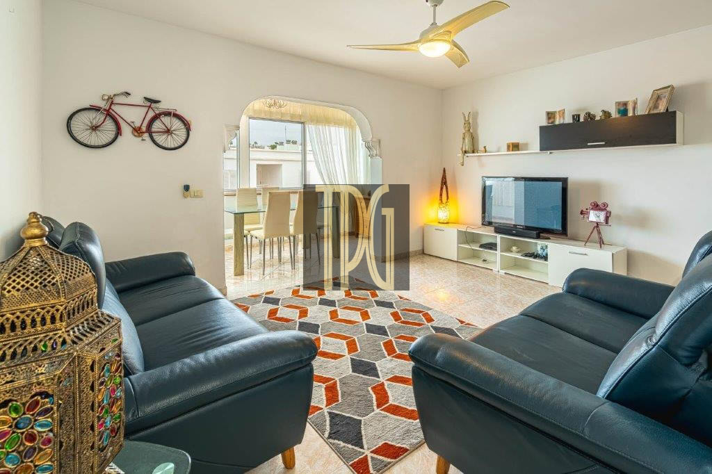 Living room with two blue leather couches facing a TV on a white console and a colorful geometric rug in a bright open space with an arched doorway to the dining area.