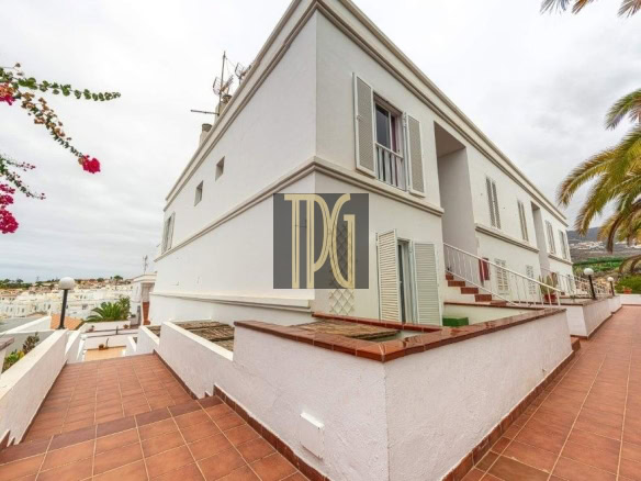 White two-story building with shutters, a tiled terrace, and an exterior staircase along a sunny coastal walkway of red tiles.