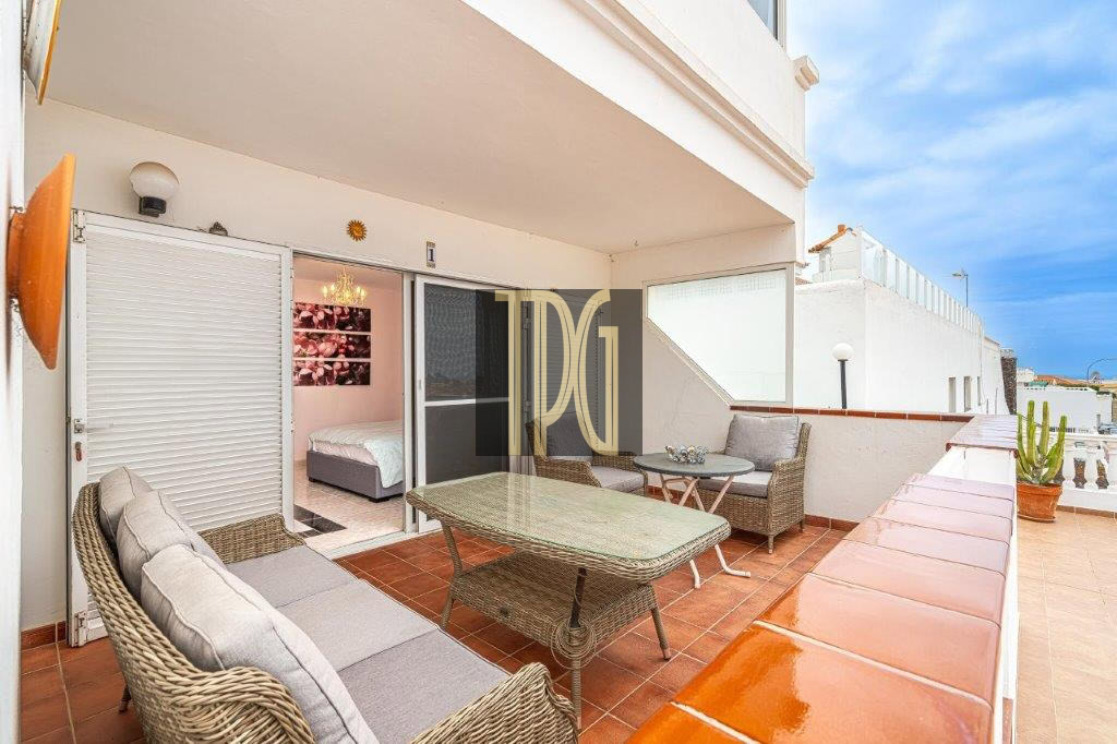 Terrace with wicker sofa set and glass table, overlooking a sunny white balcony with a potted cactus nearby and a bedroom visible through sliding doors