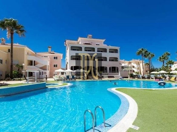 Resort pool area with bright blue water, palm trees, and pastel-colored buildings surrounding a sunny courtyard.