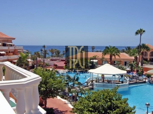Resort pool area with palm trees, pink-tiled buildings, and a view of the ocean in the distance under a clear blue sky