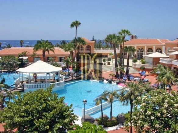 Resort pool area with palm trees, lounge chairs, and orange-roofed buildings; ocean visible in the background.2