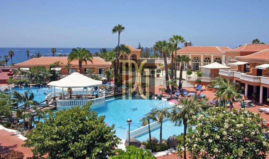 Resort pool area with palm trees, lounge chairs, and orange-roofed buildings; ocean visible in the background.2