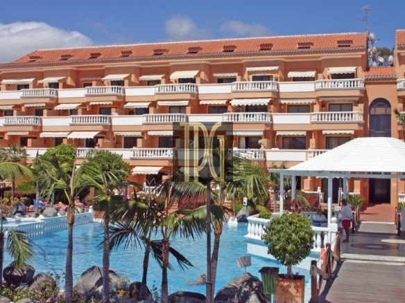 Orange multi-story resort with white balconies, a blue swimming pool in the foreground, and palm trees around a wooden walkway and gazebo site.