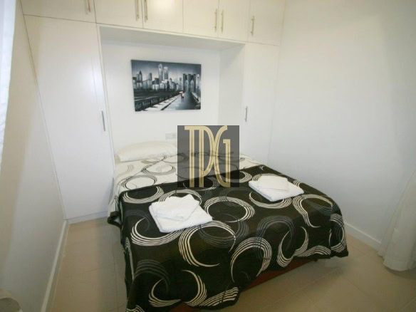 Small white bedroom with built-in wardrobes surrounding a neatly made double bed, black and white swirl-patterned bedspread, and two folded towels on the bed with a cityscape wall art above it.