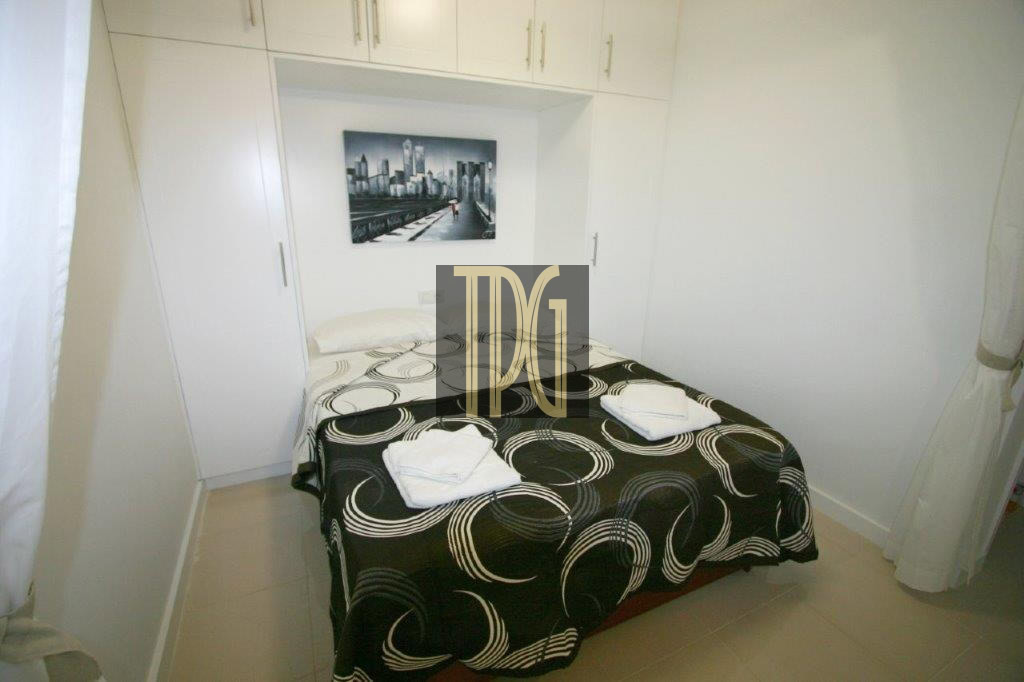 Small white bedroom with built-in wardrobes surrounding a neatly made double bed, black and white swirl-patterned bedspread, and two folded towels on the bed with a cityscape wall art above it.