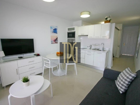 Modern open-plan living area with a white kitchenette, round dining table, TV on a white cabinet, and a gray sofa with a plaid cushion in a bright, minimal space.