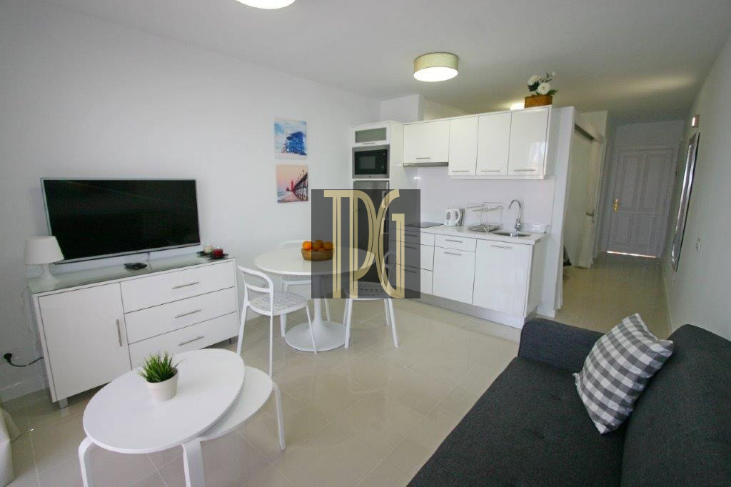 Modern open-plan living area with a white kitchenette, round dining table, TV on a white cabinet, and a gray sofa with a plaid cushion in a bright, minimal space.