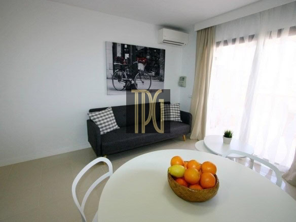 Bright living room with a dark sofa and plaid cushions, round white dining table, and a basket of oranges on the table near large windows with sheer curtains.