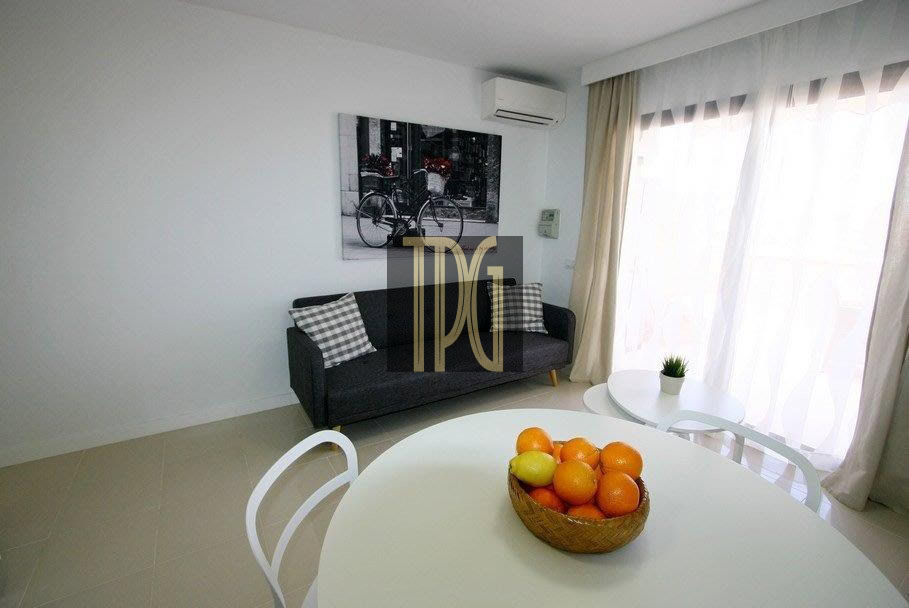Bright living room with a dark sofa and plaid cushions, round white dining table, and a basket of oranges on the table near large windows with sheer curtains.