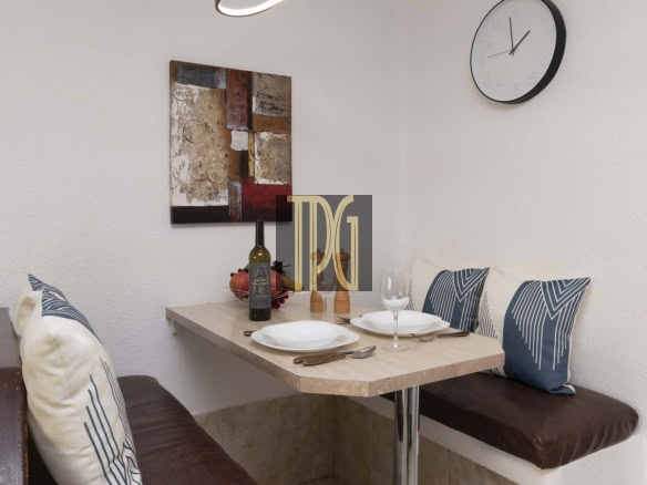 Cozy corner dining nook with a small light wood table set for two, wine bottle, two goblets, and decorative pillows against white walls.