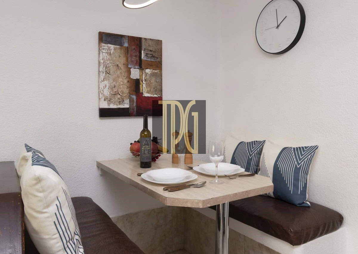 Cozy corner dining nook with a small light wood table set for two, wine bottle, two goblets, and decorative pillows against white walls.