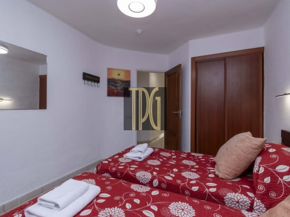 Twin bedroom with red floral bedspreads and folded towels on each bed, beige pillows, a wooden wardrobe and door, and a wall mirror.