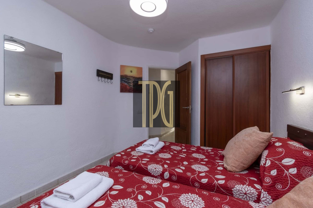 Twin bedroom with red floral bedspreads and folded towels on each bed, beige pillows, a wooden wardrobe and door, and a wall mirror.