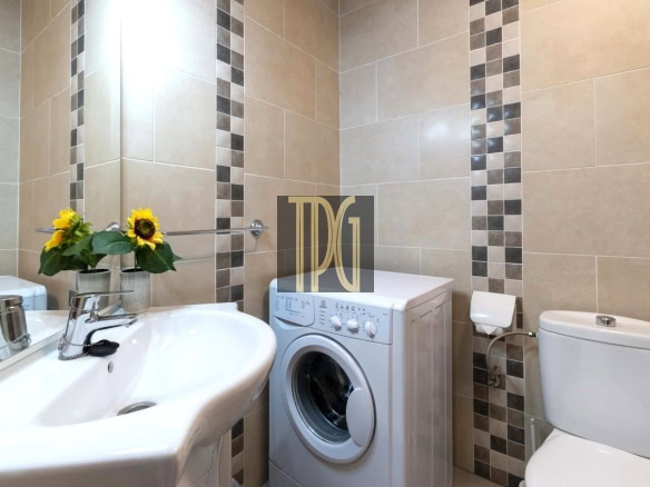 Beige tiled bathroom with a front-loading washing machine between a sink and a toilet; yellow sunflowers sit on the sink edge.