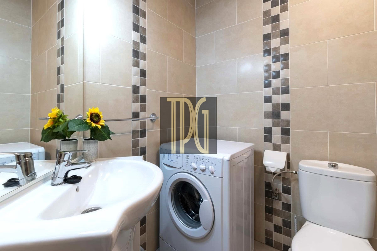 Beige tiled bathroom with a front-loading washing machine between a sink and a toilet; yellow sunflowers sit on the sink edge.