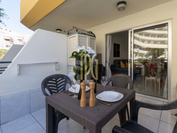 Outdoor balcony dining table set with plates, glasses, cutlery, a wine bottle, salt and pepper mills, and a floral centerpiece; sliding glass door to living room.