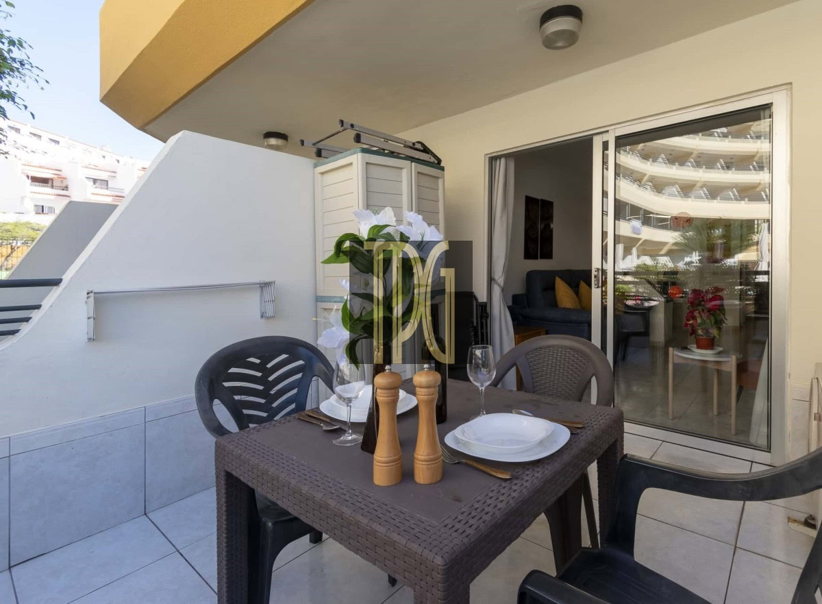 Outdoor balcony dining table set with plates, glasses, cutlery, a wine bottle, salt and pepper mills, and a floral centerpiece; sliding glass door to living room.