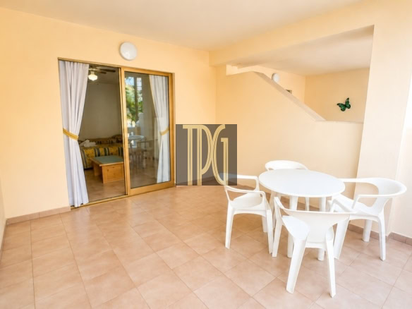 Outdoor patio with beige walls and tiled floor, a white round table and four white chairs, and a sliding glass door to an interior bedroom.