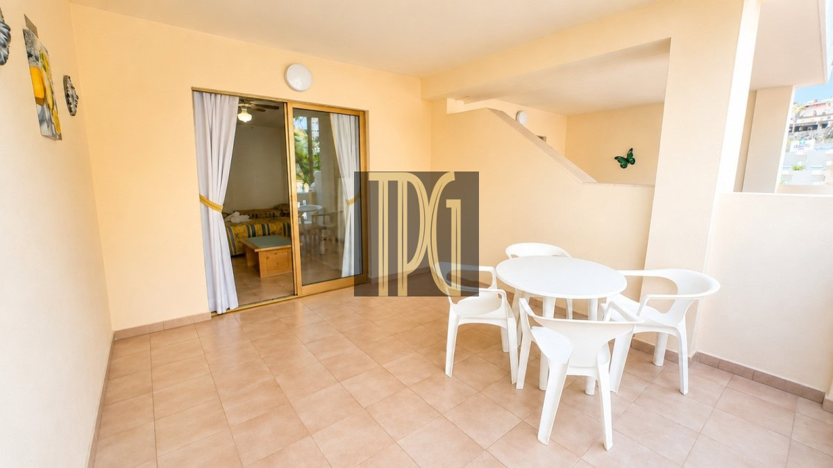 Outdoor patio with beige walls and tiled floor, a white round table and four white chairs, and a sliding glass door to an interior bedroom.