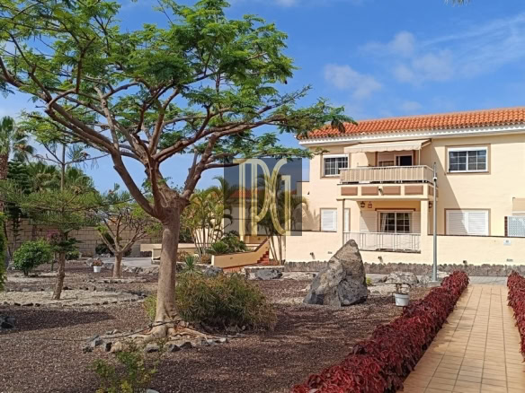 Two-story beige apartment building with balconies and a red-tiled roof, viewed from a desert-style yard with a large tree, palms, and a tiled walkway.