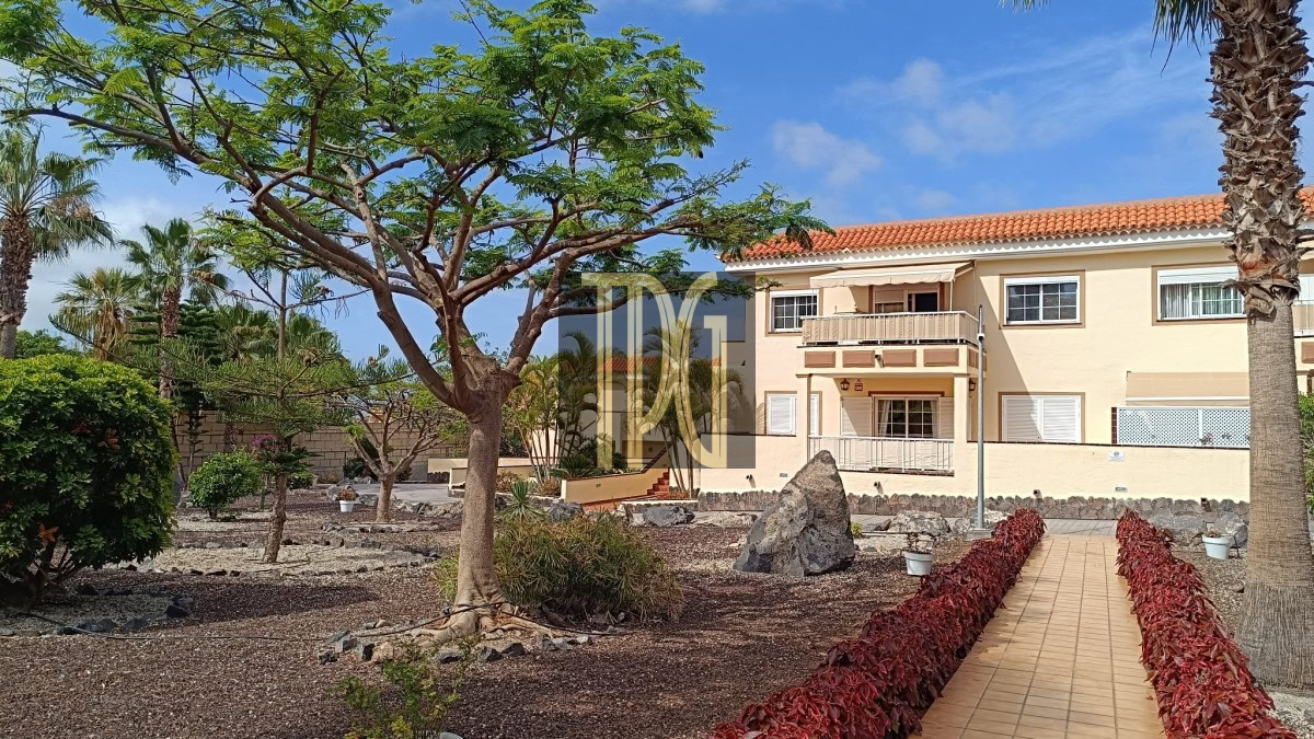 Two-story beige apartment building with balconies and a red-tiled roof, viewed from a desert-style yard with a large tree, palms, and a tiled walkway.