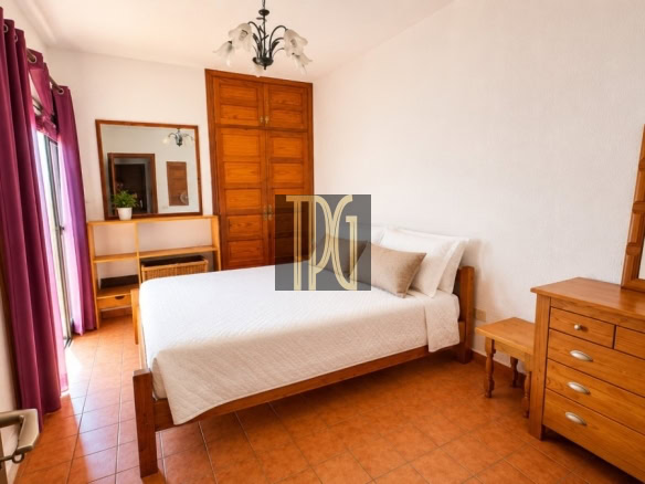 Wooden double bed in a bright bedroom with a white quilt, flanked by warm-toned dressers and a large wooden wardrobe; purple curtains frame a door to the left.